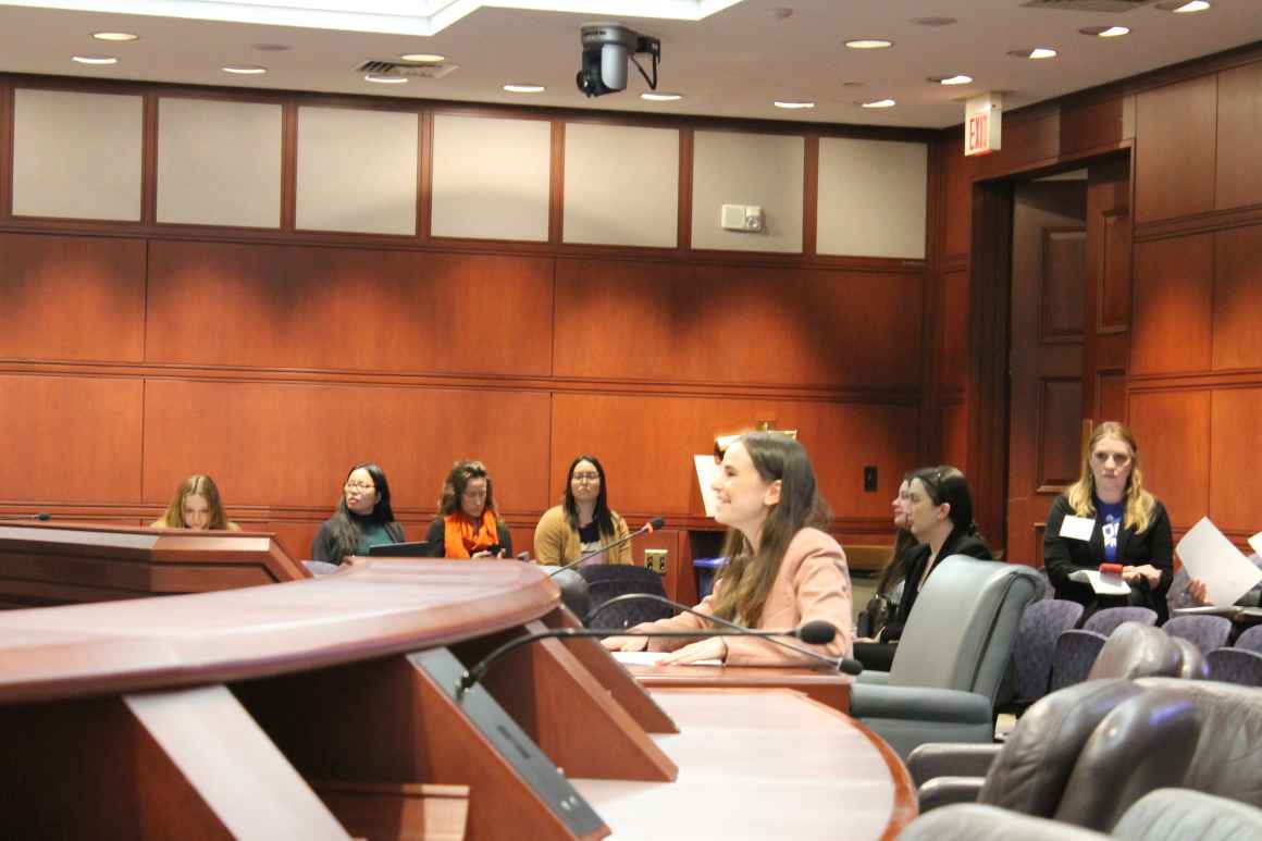 Jess, a white woman, wearing a pink blazer is testifying at the Capitol.