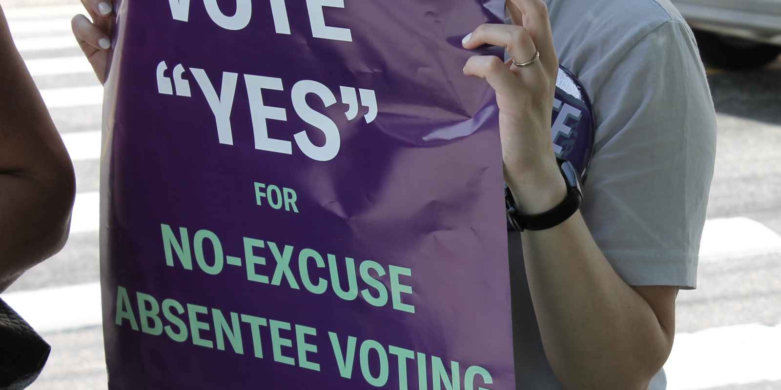 A woman, who is mostly out of frame, is holding a purple sign that says "VOTE "YES" FOR NO-EXCUSE ABSENTEE VOTING" in white and teal text.