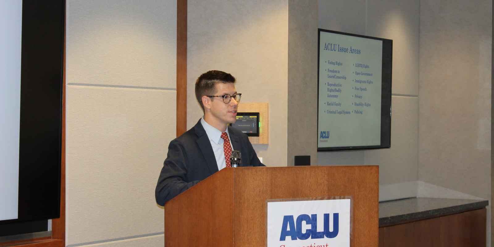 A white man with dark hair and glasses stands behind a wooden podium that has the ACLU-CT logo on it.