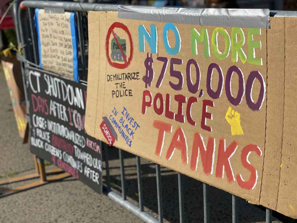 A cardboard sign taped to a fence outside of a Stamford Connecticut protest. The sign says: "No more $750,000 police tanks," "demilitarize the police," and "invest in our communities." It has a drawing of a raised fist, and of a tank crossed out.