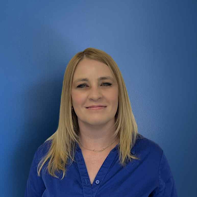 Amber Vlangas, a white woman with blonde hair wearing a blue button up shirt, looking at the camera with a smile.