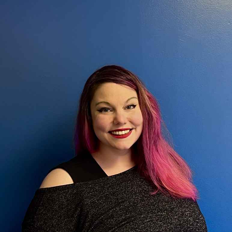 A white woman with brown hair that's dyed pink midway to the end smiles at the camera in front a blue background with a People Not Prisons tshirt and blazer on.