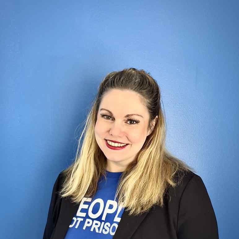 A white woman with brown hair that's bleached midway to the end smiles at the camera in front a blue background with a People Not Prisons tshirt and blazer on.