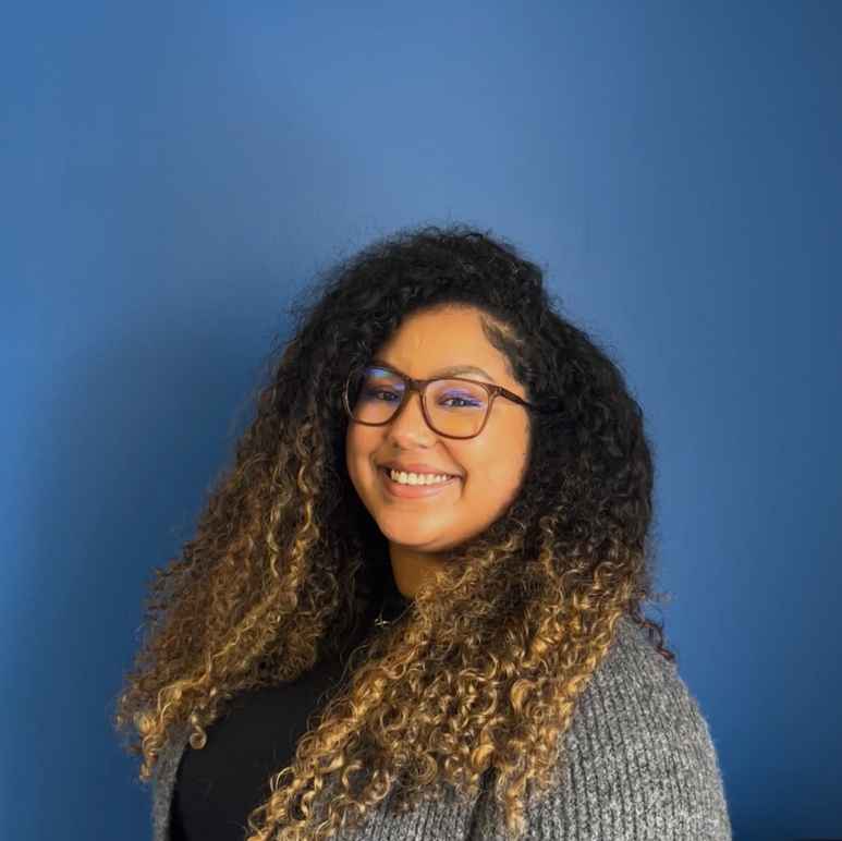 An Afro-Bori woman with curly hair, her roots are dark and her ends are bleached, in a grey cardigan smiles at the camera in front of a blue wall.