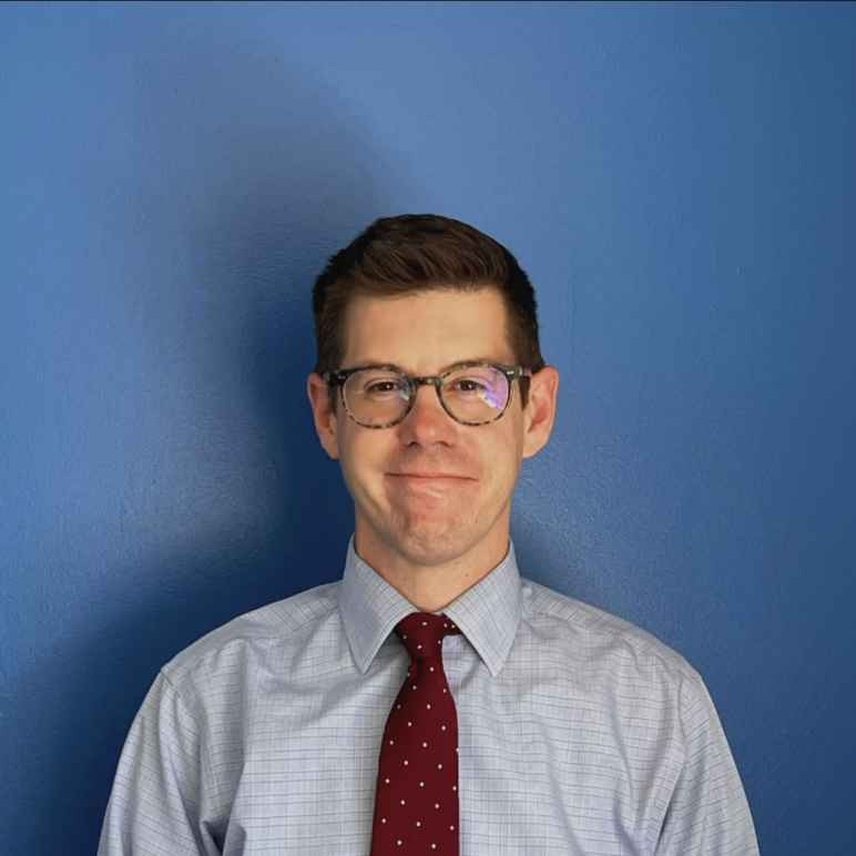 David McGuire, a white man with short brown hair wearing a light blue dress shirt with a red tie and brown glasses, looking at the camera with a smile.