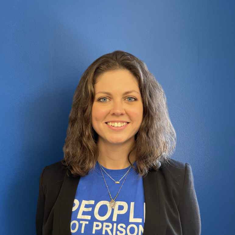 A white woman with light brown hair to her shoulders smiles at the camera in a blue People not Prisons t-shirt and blazer combo, standing in front of a blue wall.
