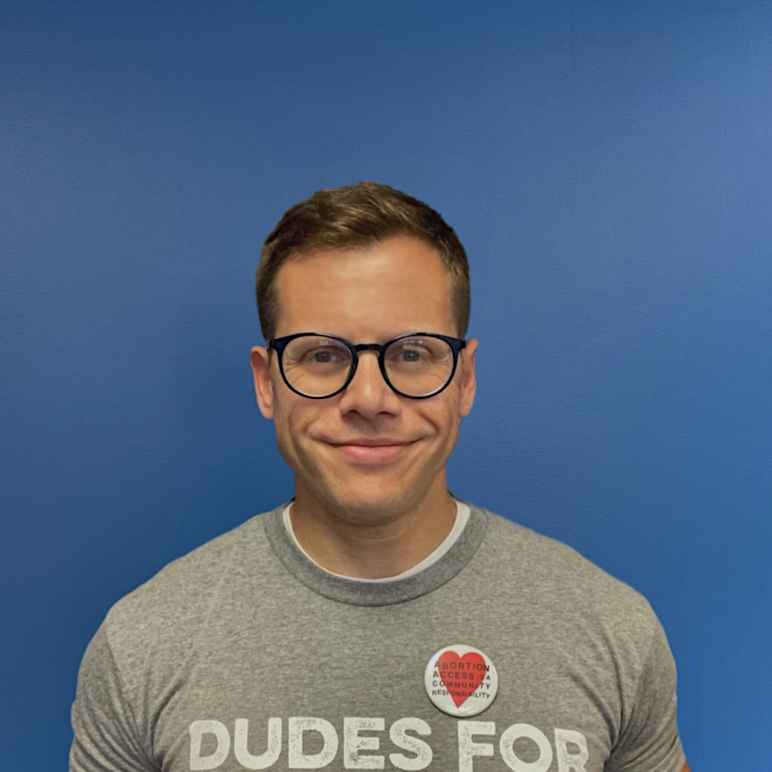 Gus Marks-Hamilton, a white man with black round glasses, smiles at the camera in front a blue wall with a gray shirt that says "Dudes for Abortions."