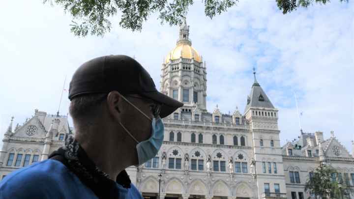 ACLUCT Smart Justice leader Sean Sellars is in the foreground, wearing a mask and baseball hat, looking toward the CT capitol building. In the background is the Connecticut capitol building and a blue sky.