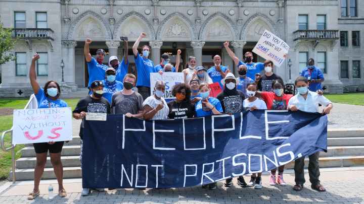 A group of Smart Justice leaders, advocates, and legislators, all wearing masks, stand behind the ACLU-CT's blue "People Not Prisons" banner outside of the Connecticut state capitol building.