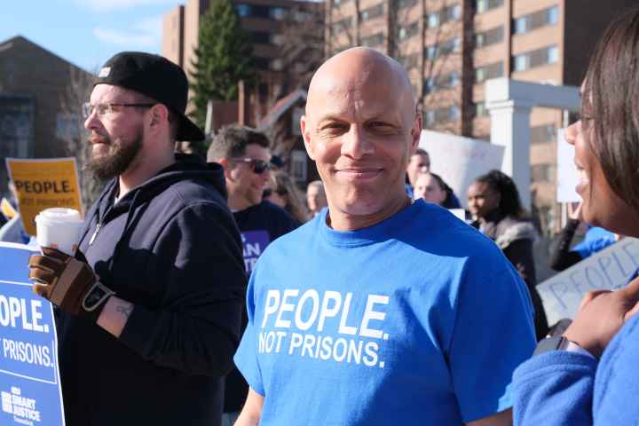 ACLUCT smart justice leader Brian Sullivan smiles at the camera. He's wearing a blue people not prisons shirt. Behind him is a crowd of people at a protest in Feb. 2020.