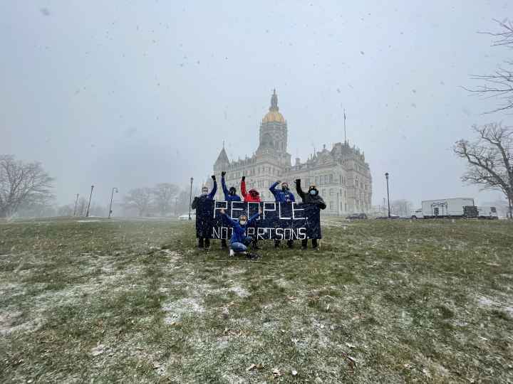 A photo of the Connecticut State Capitol and ACLU-CT Smart Justice leaders with a blue "people not prisons banner." Foreground: capitol lawn, dusted w snow. Middleground: 6 leaders, fists up, hold the banner. Background: snowy sky and capitol building
