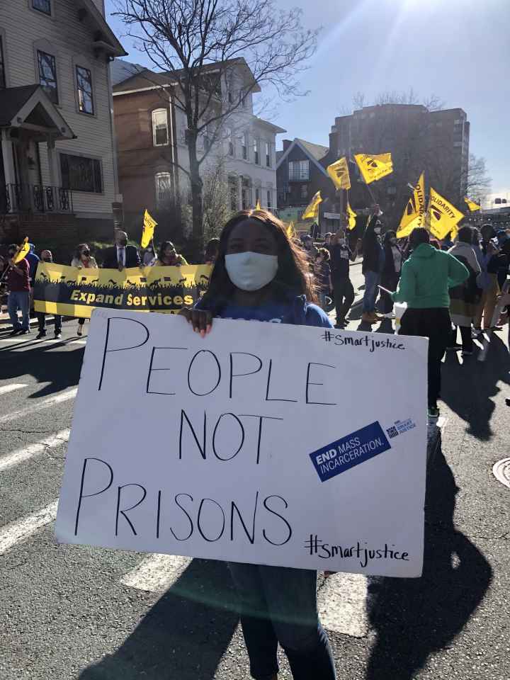 Smart Justice leader Shelby Henderson holds a white sign that says "PEOPLE NOT PRISONS" in blue ink. Behind her is a crowd waving yellow flags and carrying a banner. They are on a street in Hartford, marching.