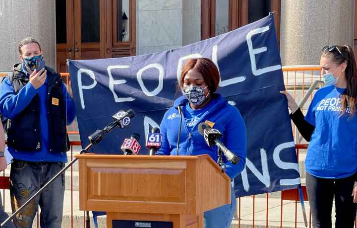 Smart Justice leader shelby Henderson, in a blue Smart Justice zip up and mask, speaks at a podium in front of the CT state capitol. Behind her is a blue people not prisons banner held by two other Smart Justice leaders.