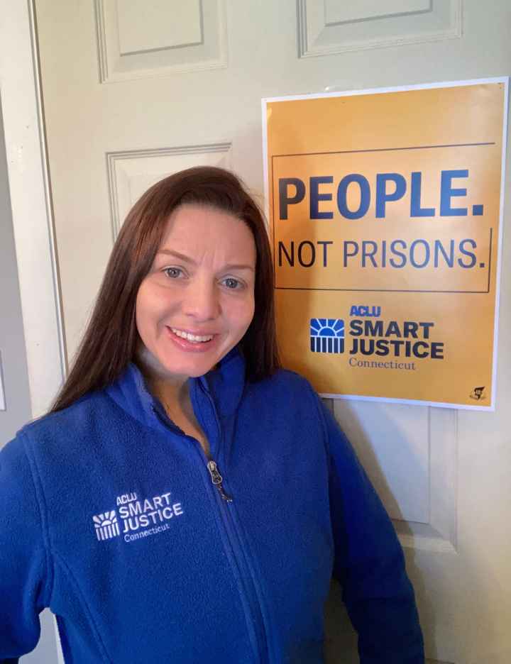 ACLU-CT Smart Justice leader Tracie Bernardi stands in front of a yellow Smart Justice "People Not Prisons" poster. She is wearing a blue ACLU of Connecticut Smart Justice zip up sweatshirt, standing, and smiling.