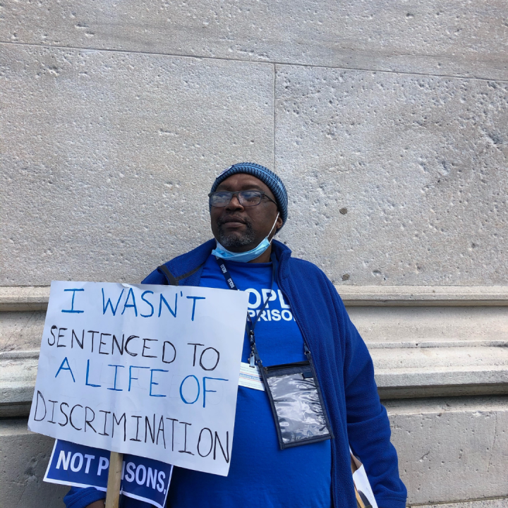 ACLUCT senior policy organizer Anderson Curtis stands with a white sign that says "I wasn't sentenced to a lifetime of discrimination." He wears a blue people not prisons shirt and sweatshirt and blue hat, glasses. He is leaning on a gray stone building