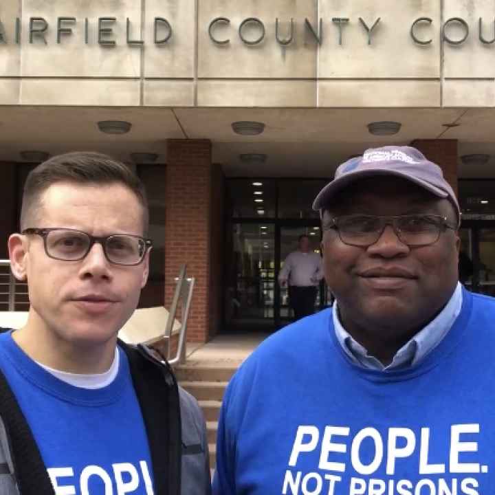 ACLU-CT Gus Marks-Hamilton and Anderson Curtis in blue "people, not prisons" shirts, stand outside of the Fairfield County Courthouse