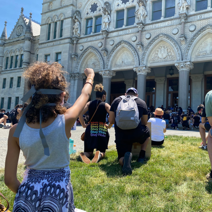A crowd of people, spaced apart for physical distancing, take a knee in front of the Connecticut capitol building. A person in the foreground, hair up in a ponytail, holds her fist up.