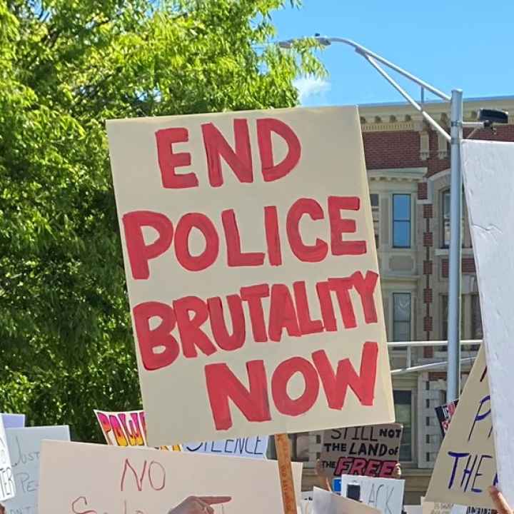 A white sign with red writing, surrounded by other protest signs and against a backdrop of trees. The sign with red writing says: END POLICE BRUTALITY NOW