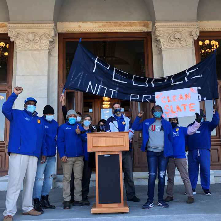 ACLU-CT Smart Justice leaders stand with "people not prisons" banner and "Clean Slate" sign outside of the CT State Capitol. They are behind a podium, and wearing blue "people not prisons" shirts. Many have fists up in power salutes.