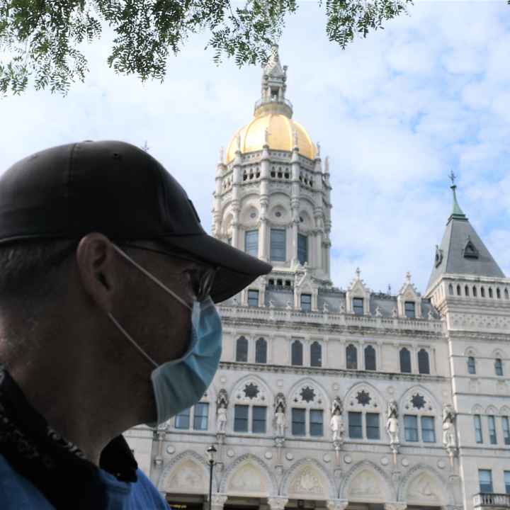 ACLUCT Smart Justice leader Sean Sellars is in the foreground, wearing a mask and baseball hat, looking toward the CT capitol building. In the background is the Connecticut capitol building and a blue sky.