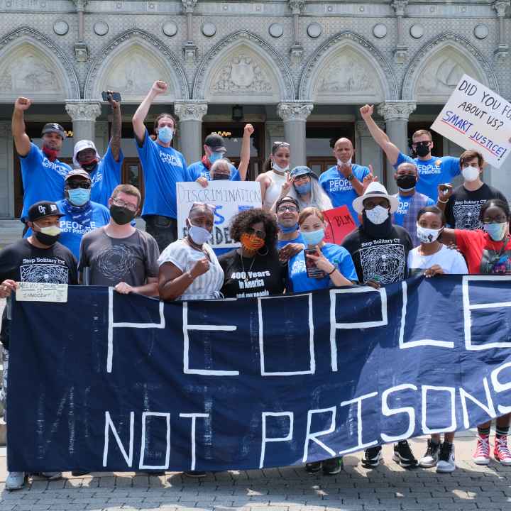 A group of Smart Justice leaders, advocates, and legislators, all wearing masks, stand behind the ACLU-CT's blue "People Not Prisons" banner outside of the Connecticut state capitol building.