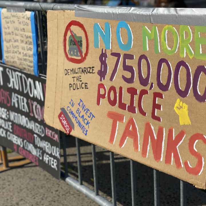 A cardboard sign taped to a fence outside of a Stamford Connecticut protest. The sign says: "No more $750,000 police tanks," "demilitarize the police," and "invest in our communities." It has a drawing of a raised fist, and of a tank crossed out.