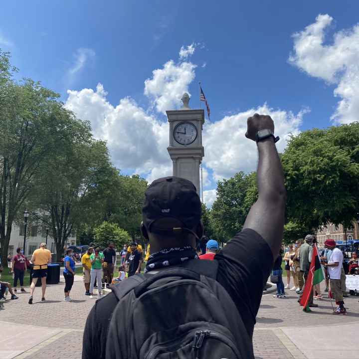 A crowd of people is gathered around the clock tower in Waterbury, Connecticut. The sky is blue and it is sunny. A Black man is in the foreground, back to the camera, with fist raised. He is wearing a backpack. 