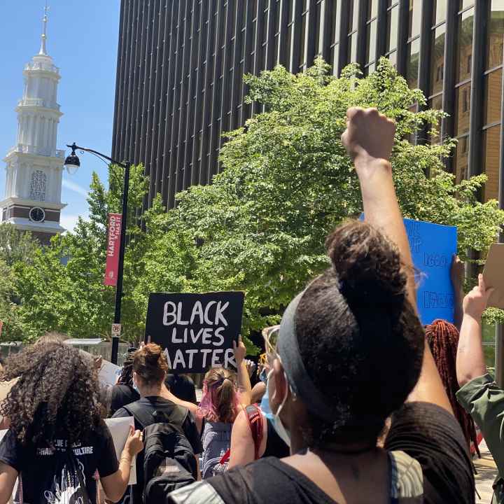 Protesters march in Hartford to call for the state to value Black lives. In the foreground, a person has their fist up. In the background, a Black Lives Matter sign
