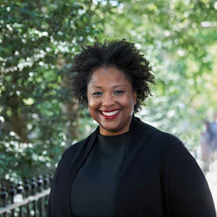 Deborah Archer, national ACLU president, is in the foreground. She is wearing a black shirt and blazer and is smiling, looking directly at the camera. Behind her is a leafy park. 
