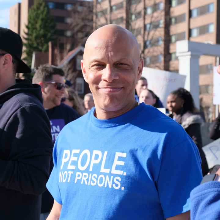 ACLUCT smart justice leader Brian Sullivan smiles at the camera. He's wearing a blue people not prisons shirt. Behind him is a crowd of people at a protest in Feb. 2020.
