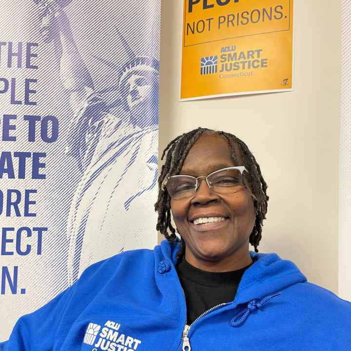 ACLUCT smart justice leader Terri Ricks sits, smiling and looking straight at the camera. She is wearing a blue ACLU of Connecticut Smart justice hoodie and glasses. Behind her is a yellow people not prisons poster.
