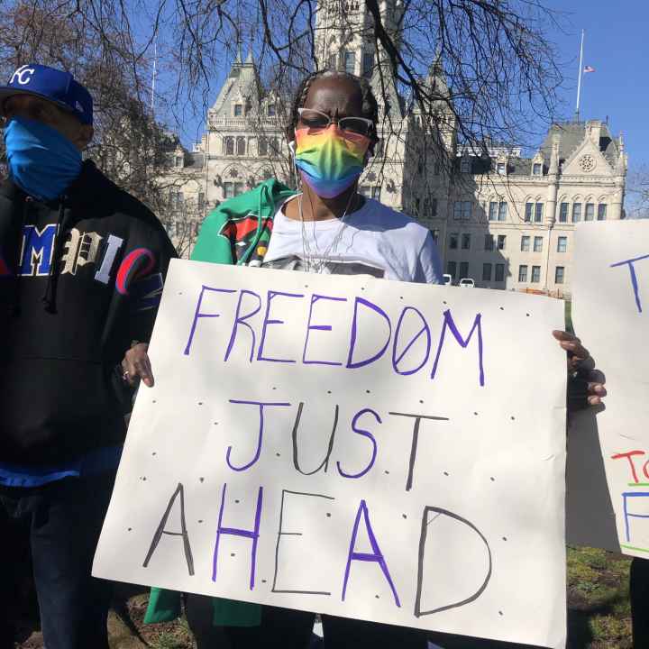 ACLU-CT Smart Justice leader Terri Ricks stands in front of the CT State Capitol Building. She is wearing sunglasses and a rainbow face mask, and she holds a white sign that says, in blue letters, "FREEDOM JUST AHEAD."