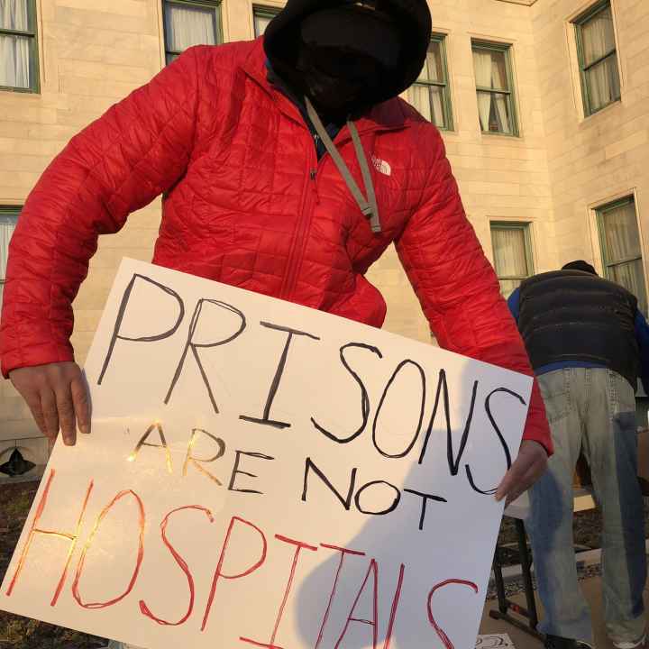 A person in a red coat, black facemask, and black hood holds a sign that says "Prisons are not hospitals" in front of the Connecticut State Capitol building in December 2020.