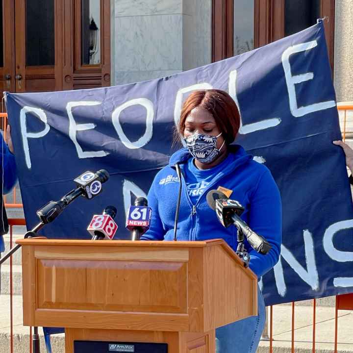 Smart Justice leader shelby Henderson, in a blue Smart Justice zip up and mask, speaks at a podium in front of the CT state capitol. Behind her is a blue people not prisons banner held by two other Smart Justice leaders.