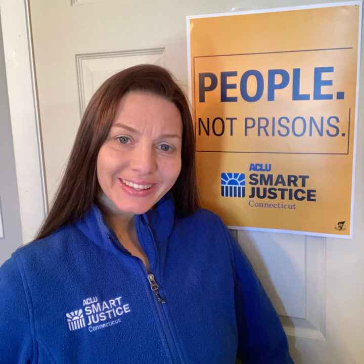 ACLU-CT Smart Justice leader Tracie Bernardi stands in front of a yellow Smart Justice "People Not Prisons" poster. She is wearing a blue ACLU of Connecticut Smart Justice zip up sweatshirt, standing, and smiling.