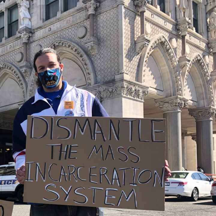 ACLUCT smart justice leader Will stands with a cardboard sign that says, in black and white, "dismantle the mass incarceration system." Behind him is the CT Capitol. he is wearing a mask and a sweatshirt, and yellow people not prison pin
