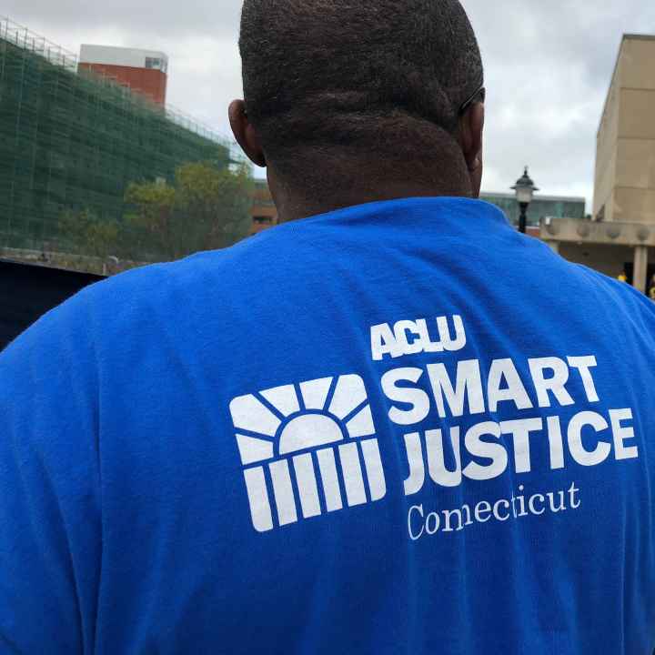 From behind, a man stands in a blue ACLU of Connecticut Smart Justice shirt