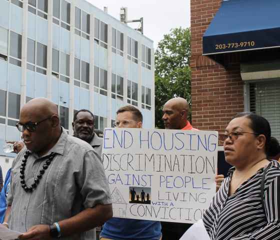 A crowd stands in front of a rental management building with signs to advocate for equitable housing. Gus, our campaign manager, is in the center in focus with a sign that says "end housing discrimination against people with a record of conviction."